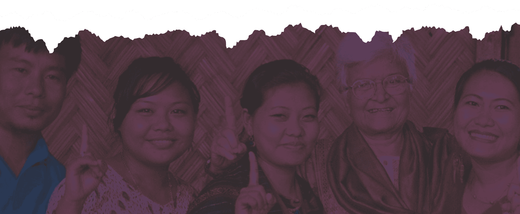 Three indigenous women, two young and one elderly, smiling and raising a finger together.